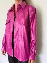 Camisa magenta de cuero sintético, de corte recto, con cuello clásico, cierre frontal con botones a presión, dos bolsillos de parche con solapa en el pecho y mangas largas con puños abotonados.