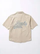 Camisa de manga corta color crudo, confeccionada en gabardina, con cuello camisero y botones frontales. Presenta un bolsillo de parche en el pecho y bordado en la espalda.