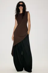 Vestido corto de color marrón, con cuello redondo y diseño sin mangas. Presenta un bajo asimétrico con un lateral más largo y cierre en la espalda con abertura y botón.