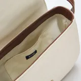 Bolso bandolera de diseño estructurado, confeccionado en lona color beige con detalles de cuero sintético marrón en los bordes y la solapa frontal. Cuenta con una hebilla decorativa metálica, costuras a la vista en color blanco y correa ajustable.