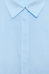 Camisa amplia confeccionada en hilatura con lyocell y ramio, color celeste, con cuello solapa, manga por debajo del codo, detalle de pliegue en la espalda y cierre frontal con botones ocultos por solapa.