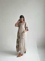 Vestido largo beige de lino con diseño midi holgado, cuello redondo, mangas cortas con vuelta y un gran estampado lateral de flores y hojas en tonos marrones y verdes. Cuenta con un bolsillo frontal aplicado con detalles deshilachados.