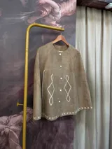 Capa de cuero genuino con textura de reptil, color beige, con cierre frontal de botones y detalles de pasamanería bordada a mano en color blanco. Presenta un diseño de rombos en el frente y costuras decorativas en forma de cruz en el ruedo.