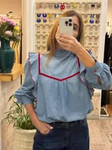 Blusa de jean fino con cuello alto, mangas largas con volumen y volados decorativos en los hombros. Presenta detalles de ribetes en terciopelo azul oscuro en el frente y los volados.