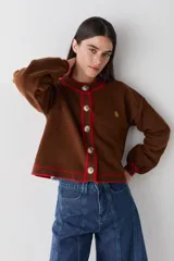 Cardigan de punto color marrón con detalles de ribete en color verde oliva en cuello, puños, ruedo y tapeta de botones. Presenta cierre frontal con botones grandes y un pequeño bordado en el pecho.