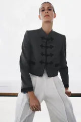 Blazer de corte cropped confeccionado en tejido de lana gris, con cuello redondo y cierre frontal mediante alamares decorativos. Presenta bolsillos delanteros con solapa y puños a contraste en color negro.