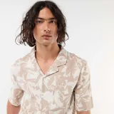 Camisa de manga corta color beige con estampado abstracto de hojas blancas, cuello camisero abierto y cierre frontal de botones.