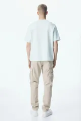 Pantalón cargo color beige de corte slim fit, confeccionado en tejido de algodón. Cuenta con cintura elástica ajustable con cordón, bolsillos al bies, bolsillos laterales y traseros con solapa y botones a presión ocultos, costuras y pinzas en las rodillas.