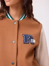 Campera estilo varsity con cuerpo en color marrón y mangas en tono beige. Presenta cierre frontal con botones a presión, cuello, puños y ruedo con franjas tejidas en verde y blanco. Incluye un parche con la letra R en el pecho y detalles bordados en las mangas.