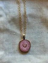 Collar de cadena fina dorada con un dije circular que presenta un centro de resina en tono rosado con efecto marmolado y un pequeño símbolo de media luna en el centro.