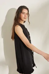 Vestido corto de punto sin mangas, color negro, con cuello redondo y cierre frontal decorado con botones y tiras colgantes. Presenta terminaciones con volados en el ruedo.