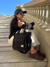 Cartera tipo tote bag negra de lona, con bolsillo frontal con solapa, doble asa de mano y correa ajustable para el hombro.