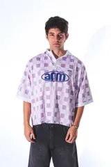 Camiseta tipo polo de estilo deportivo, con diseño a cuadros en tonos blanco y lila. Presenta cuello polo, mangas cortas y estampa gráfica circular en el frente con la inscripción 'anm'.