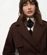 Trench coat largo color marrón chocolate, confeccionado en tejido tipo paño, con solapas amplias, cierre frontal cruzado con cinturón de la misma tela para anudar y detalles de trabillas en los hombros y puños.