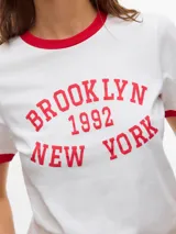 Remera blanca de manga corta con cuello redondo y ribetes rojos. Estampa frontal con la palabra "BROOKLYN" y "NEW YORK" en rojo.