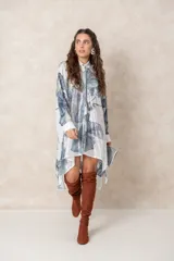 Vestido tipo túnica de corte irregular, con cierre frontal de botones y cuello camisero. Presenta un estampado botánico en tonos azules y verdes sobre fondo blanco, con detalles bordados brillantes en la parte frontal.