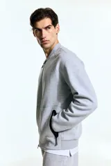 Campera bomber deportiva gris melange, de corte estándar, con cuello elevado corto, cierre bidireccional y mangas raglán. Posee bolsillos delanteros con cierre y costuras selladas, además de elástico fino en puños y bajo.