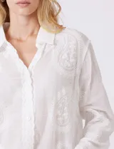 Camisa de gasa ligera y traslúcida, color blanco, con intrincados bordados búlgaros y motivos botánicos distribuidos en toda la prenda. Presenta una guarda bordada que recorre la línea de botones, el ruedo y los puños. Tiene cuello con solapa y abertura central con botones. La silueta es amplia y relajada, con mangas largas y un canesú en la espalda para una caída impecable.