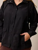 Campera de corte recto y estilo funcional, color beige, con capucha integrada, cierre frontal con botones a presión y bolsillos delanteros amplios. Presenta un diseño minimalista con cordones ajustables en la capucha.