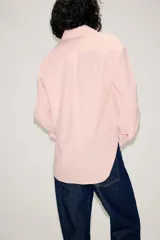 Camisa de corte oversize color rosa claro con diseño de rayas verticales sutiles. Presenta cuello solapa desmontable, cierre frontal con botones ocultos bajo solapa, mangas largas con puños y bajo asimétrico redondeado. Incluye dos bolsillos frontales amplios.
