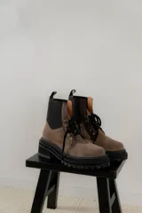 Borcego de gamuza color chocolate con paneles laterales elásticos negros y suela track dentada. Presenta sistema de cordones con ganchos metálicos y tirador en la parte trasera.