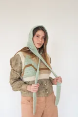 Capucha tejida a mano en lana merino con diseño de rayas horizontales en color mostaza y beige. Presenta una forma envolvente con lazos largos para anudar bajo el cuello, funcionando como un accesorio independiente de abrigo.