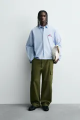 Camisa de algodón de corte regular, color celeste con finas rayas blancas verticales. Presenta un bordado en color rojo en el pecho con la frase "Café du Matin Doux".