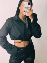 Camisa cropped negra con cuello clásico, mangas largas con puños abotonados y cierre frontal con botones. Presenta detalle de tachas plateadas en el cuello.