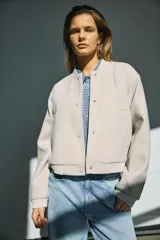 Campera bomber de corte holgado en color beige, con cuello redondo tipo baseball, cierre frontal con botones a presión, bolsillos laterales y puños y ruedo elastizados.