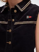 Chaleco de pana negro con cierre frontal de botones metálicos dorados. Presenta detalles de costuras en contraste en el cuello, cartera y canesú, e incluye un pequeño parche con forma de bandera en el pecho.