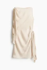 Vestido corto sin mangas de color beige con un leve brillo, corte entallado, elegante escote bote y tiras anchas para atar en un hombro y en el lado opuesto de la cintura. Cuenta con cierre oculto en un lateral.