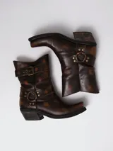 Botas estilo cowboy de cuero en tono marrón con efecto desgastado. Presentan un diseño de caña media con detalles de correas, argollas metálicas y tachas decorativas. Cuentan con un taco cuadrado de altura media y punta ligeramente cuadrada.