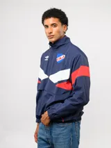 Buzo deportivo de nylon con medio cierre frontal y cuello alto. Presenta un diseño de bloques de color en azul, blanco y bordó, con el escudo del Club Nacional de Football y el logo de Umbro en el pecho.