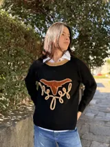 Sweater de punto fino color verde, con cuello redondo en contraste color beige. Presenta un diseño frontal con una figura de búfalo en cuero y detalles bordados a mano en color beige.