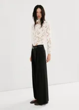 Blusa de encaje blanco con diseño floral, cuello alto y mangas largas con hombreras. Presenta un corte ajustado y acabado delicado.
