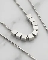 Collar doble de acero con cadenas finas. La cadena superior presenta varios cubos pequeños alineados, mientras que la cadena inferior cuenta con un dije central en forma de cubo.