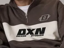 Buzo de cuello alto con medio cierre y diseño en bloques de color marrón y una franja blanca ancha en el pecho. Cuenta con estampado tipográfico de la marca "DXN Duxon Studios" en color negro sobre la franja blanca y un logotipo pequeño en el pecho.