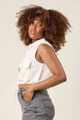 Chaleco cropped color crema, confeccionado en lino, con cuello camisero, cierre frontal con botones, dos bolsillos con solapa en el pecho y lazo ajustable en la cintura.