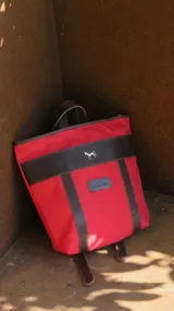 Mochila de lona roja con detalles y correas de cuero marrón, cierre superior y aplique metálico con forma de caballo.