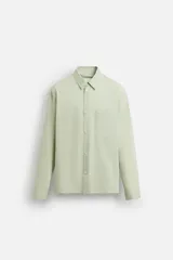 Camisa de hombre de corte relajado (relaxed fit) en color verde menta pálido, confeccionada en tejido con textura irregular acanalada. Presenta cuello solapa, manga larga con puño abotonado, bolsillo plastrón en el pecho y bajo asimétrico con aberturas laterales.