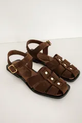 Sandalias planas de gamuza marrón, estilo pescador, con tiras entrelazadas, tachas doradas y tira ajustable con hebilla en el tobillo.