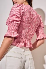 Blusa de algodón con diseño a rayas verticales en tonos rosa y blanco. Presenta un cuerpo con fruncido elástico (smock), cuello alto con volados y mangas tres cuartos abullonadas con terminación en volado.