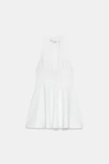 Vestido mini de color blanco con cuello halter, escote en pico y falda con vuelo tipo capa. Presenta cierre con cremallera oculta en la espalda y lazada en el cuello.