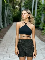 Falda corta cruzada de tejido de punto con lurex, color negro con destellos dorados. Presenta un diseño envolvente con una hebilla decorativa de madera en la cintura.