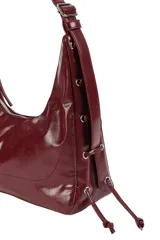Cartera tipo hobo de material sintético con acabado brillante, cierre superior con cremallera, correa ajustable y laterales con cordones decorativos.