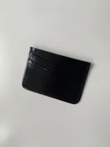 Tarjetero de cuero vacuno color negro con acabado brillante. Cuenta con cuatro ranuras para tarjetas y un bolsillo central.