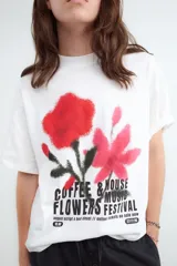 Remera de corte oversize y silueta boxy, color blanco, con cuello redondo y mangas cortas. Presenta una estampa frontal con diseño de flores en tonos rojos y rosados con efecto spray y tipografía negra.