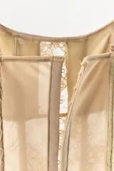 Vestido corto semitransparente tipo corsetero color beige, confeccionado con encaje y detalles de varillas interiores que estilizan la figura. Presenta escote redondo, tirantes finos ajustables y bajo asimétrico con una lazada lateral de satén.