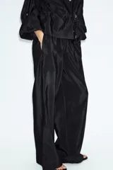 Pantalón de tiro alto color verde militar, confeccionado en tejido satinado con acabado brillante. Presenta cintura elástica fruncida, bolsillos laterales ocultos en las costuras y perneras de corte ancho.