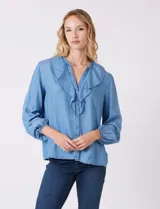 Blusa de lyocell color azul claro, con escote en V y volados en el cuello y la parte frontal. Tiene mangas largas con puños elásticos y cierre de botones en la parte delantera.